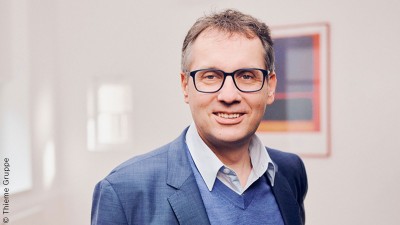 Ein älterer Mann mit blauem Anzug und Brille schaut freundlich in die Kamera. 