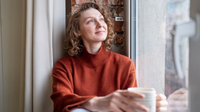 Frau mit Tasse schaut gelassen und nachdenklich aus dem Fenster