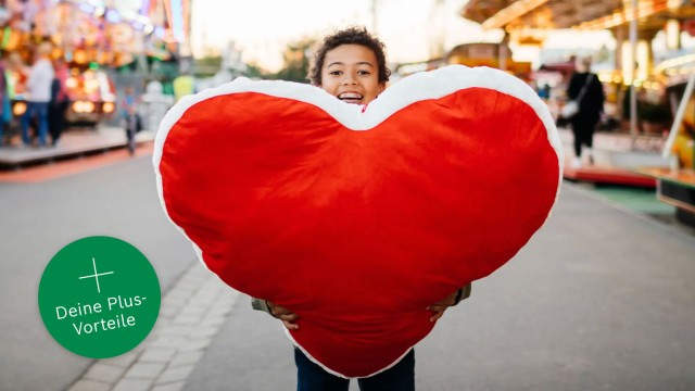 Ein Junge steht auf einem Weg in einem Jahrmarkt und hält ein großes Plüschherz. 