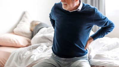 Ein älterer Mann sitzt auf dem Bett und hält sich den unteren Rücken