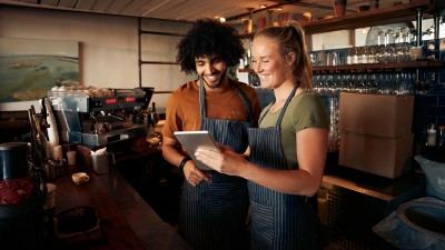 Zwei junge Menschen stehen hinter einer Bar und schauen lächelnd auf ein Tablet.