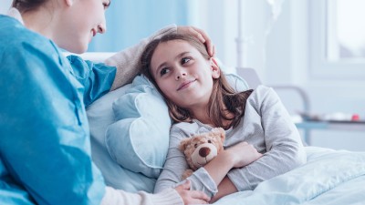 Eine Mutter sitzt in Krankenhauskleidung am Krankenbett ihrer Tochter, die einen Teddybären im Arm hält