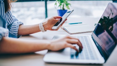 Eine Frau hält ihr Handy und tippt gleichzeitig auf der Tastatur ihres Laptops