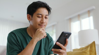 Ein junger asiatischer Mann sitzt auf dem Sofa und schaut auf sein Handy