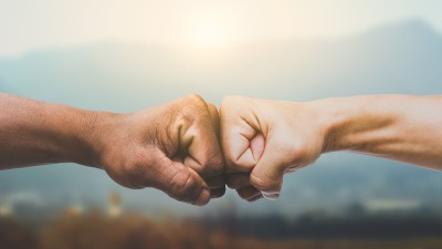 Zwei Männer geben sich einen Fauststoß, Im Hintergrund ist verschwommen Natur zu erkennen.
