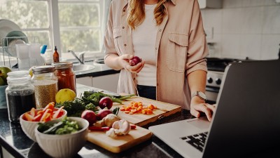 Eine junge Frau steht in der Küche vor dem Laptop und kocht mit Gemüse