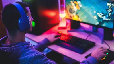 Ein männlicher Teenager mit Kopfhörern auf sitzt am Computer und spielt, das Zimmer ist in buntes Licht gehüllt.