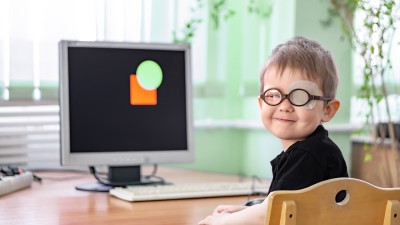 Ein kleiner Junge mit Brille und abgeklebtem Auge blickt in die Kamera, er sitzt vor einem Computerbildschirm