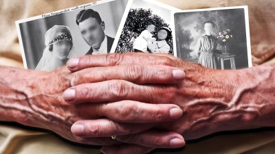 Zu sehen sind die Hände einer alten Frau, verschränkt vor dem Bauch. Sie hält Bilder mit Erinnerungen in den Händen, die Gesichter der Personen darauf sind verschwommen.