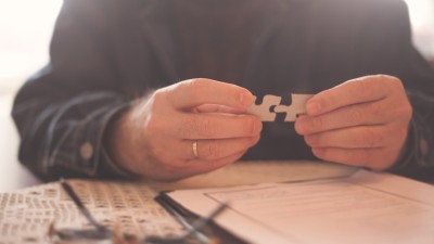 Ein alter Mann hält zwei Puzzleteile in den Händen, vor ihm liegen eine Brille und ein Blatt Papier.