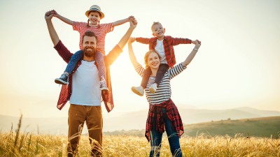 Eine junge Familie steht im Abendlicht auf einem Feld, der Vater trägt die Tochter auf den Schultern, die Mutter trägt den Sohn auf den Schultern.
