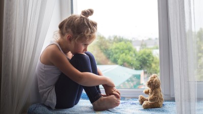 Ein kleines, blondes Mädchen sitzt vor dem Fenster, das Kinn auf die Knie gestützt und sieht traurig ihren kleinen Teddy an