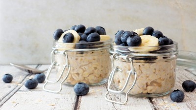 Zwei Gläser mit Overnight Oats, garniert mit frischen Blaubeeren und Bananenscheiben, stehen auf einem rustikalen Holztisch. Im Hintergrund ist ein Löffel zu sehen, und einige Blaubeeren liegen verstreut auf dem Tisch.