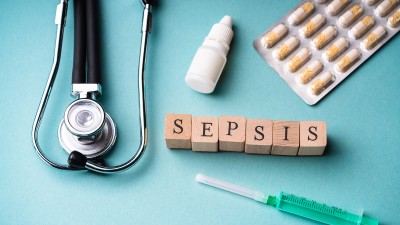 Auf hellblauem Untergrund liegen ein Stethoskop, ein Fläschchen für Augentropfen, ein Blister mit Kapseln und eine Spritze. Dazwischen ist mit kleinen Holzwürfeln das Wort „Sepsis“ in Großbuchstaben gelegt.