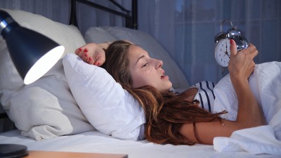 Eine junge Frau mit Schlafproblemen liegt wach im Bett und hält in den Händen einen Wecker, die Nachttischlampe neben dem Bett brennt. Sie wirkt erschöpft.