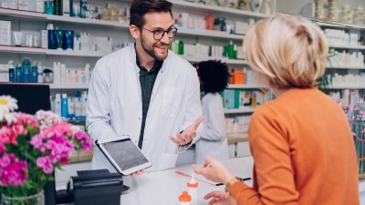 In der Apotheke berät ein Apotheker eine ältere Dame. Er hält in der Hand ein Tablet, auf dem Tresen stehen Medikamente.