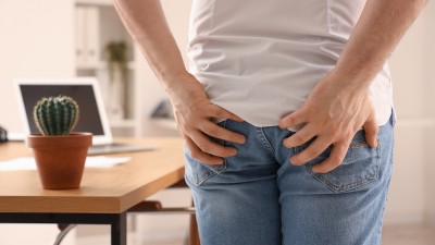 Person mit Jeans greift sich an den unteren Rücken, im Hintergrund ein Schreibtisch mit einem Kaktus.