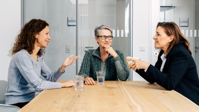 Drei Frauen mittleren Alters im Gespräch an einem Tisch mit Wassergläsern.