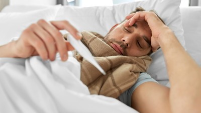 Ein Mann liegt im Bett, hält sich die Stirn und blickt auf ein Fieberthermometer.