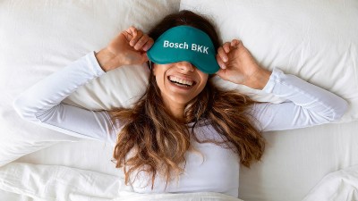 Eine glücklich aussehende Frau liegt lachend im Bett und trägt eine Schlafmaske mit dem Aufdruck "Bosch BKK". Die Bettwäsche und ihr Oberteil sind weiß.