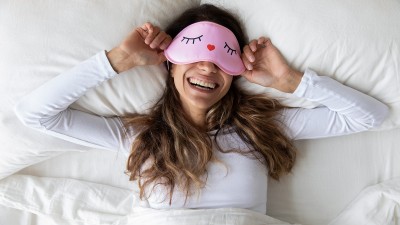 Eine glücklich aussehende Frau liegt lachend im Bett und trägt eine rosa Schlafmaske mit geschlossenen, aufgemalten Augen. Die Bettwäsche und ihr Oberteil sind weiß.