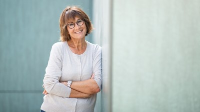 Eine Frau mittleren Alters lehnt an einer Wand und blickt in die Kamera.
