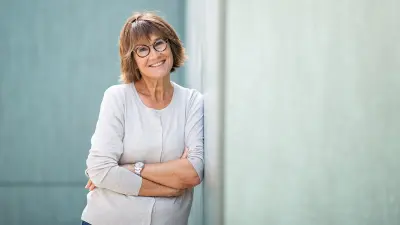 Eine Frau mittleren Alters lehnt an einer Wand und blickt in die Kamera.