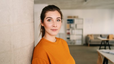 Eine junge Frau lehnt an einer Wand und blickt in die Kamera.