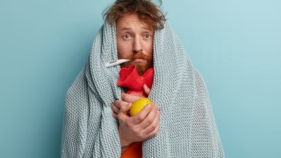 Ein Mann steht in eine Decke gewickelt vor einem hellblauen Hintergrund. Er hat ein Fieberthermometer im Mund und hält in den Händen eine Wärmflasche und eine Zitrone.