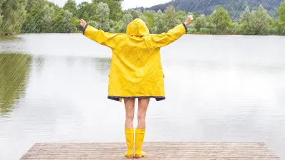 Eine Frau steht mit dem Rücken zur Kamera im Regen an einem See, sie trägt gelbe Gummistiefel und einen gelben Regemantel und streckt die Arme in die Höhe.