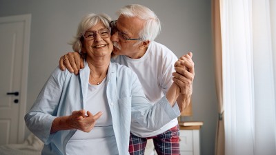 Ein Senioren-Ehepaar in einem Zimmer. Sie schaut in die Kamera, er steht hinter ihr und küsst sie auf die Wange.