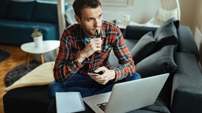 Ein Mann sitzt auf dem Sofa, vor sich Laptop und Block. Er hält ein Handy in der Hand und denkt nach.