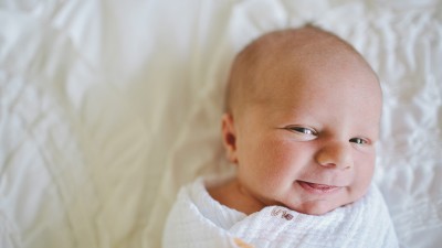 Ein Säugling liegt in eine weiße Strickdecke gewickelt auf weißem Hintergrund und lächelt in die Kamera.