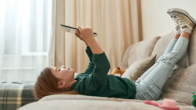 Ein Mädchen liegt auf dem Sofa, hat die Füße entspannt auf der Sofalehne abgelegt und schaut auf ein Tablet.