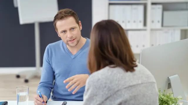 Ein Mann und eine Frau sitzen sich an einem Bürotisch gegenüber. Der Mann erklärt der Frau etwas.