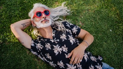 Älterer Mann mit langen weißen Haaren liegt im Graß und trägt eine rote Sonnenbrille.