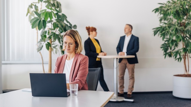 Frau am Laptop und Personen unterhalten sich im Hintergrund